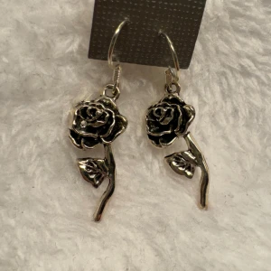 Silvriga rosformade örhängen - Säljer ett par snygga örhängen i form av rosor i silverfärg. De har en detaljrik design med både blomma och blad, perfekta för att ge din stil en romantisk touch. Krokarna är i sterling silver 