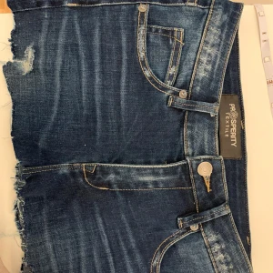 Mörkblå jeansshorts med fransig kant från Prosperity Textile - Snygga mörkblå jeansshorts med slitna detaljer och fransig kant. Klassisk femficksmodell med gylf och knapp framtill. Perfekta för sommaren och ger en avslappnad look.