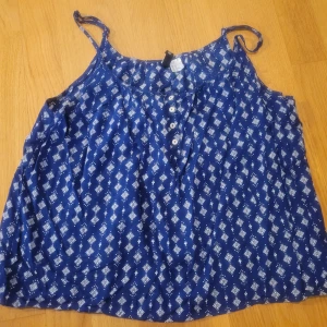 Blå mönstrad linnetopp från Divided - Säljer en blå linnetopp från Divided med vitt geometriskt mönster. Toppen har smala axelband som kan justeras och knappar framtill. Perfekt för varma dagar och enkel att matcha med jeans eller shorts.