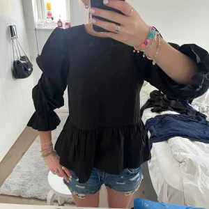 Svart blus med volang och öppen rygg - Säljer en svart blus med puffiga trekvartsärmar, volang nertill och snygg öppen rygg med knytband. Perfekt för dig som gillar trendiga detaljer och en feminin look.
