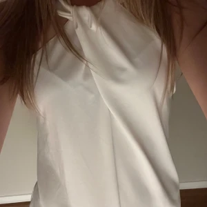 Vit halterneck topp med knyt - Säljer en stilren vit halterneck topp med knyt vid halsen. Toppen är ärmlös och har en elegant, lös passform som passar perfekt till sommaren. Materialet känns mjukt och svalt mot huden.