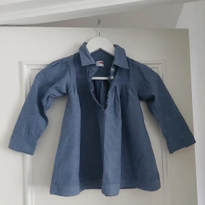 Blå jeansklänning från Name It,storlek  4 år - Supersöt blå jeansklänning från Name It med lång ärm och klassisk krage. Klänningen har knappar upptill och är i mjukt denimtyg. Perfekt för dig som gillar enkel och stilren look.