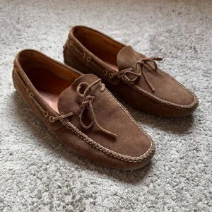Bruna mocka loafers från car shoe - Nypris på 7800kr Säljer ett par klassiska bruna loafers från Car Shoe i mjuk mocka. Skorna har dekorativ snörning framtill, ljusa sömmar och guldiga öljetter på sidorna. Yttersulan har gummidetaljer för extra grepp. Perfekta för en stilren look.