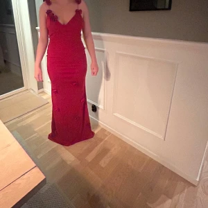 Röd balklänning med blomdetaljer - Säljer en elegant röd balklänning med smala axelband och djup rygg. Klänningen har stora volangblommor längs urringningen och på kjolen för en extra wow-effekt. Perfekt för bal eller festkvällar när du vill sticka ut. Pris kan diskuteras 