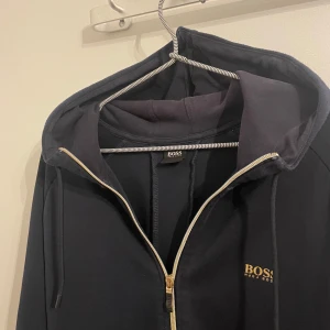 Svart hoodie med dragkedja från BOSS - Säljer en svart hoodie från BOSS med guldfärgad logga på bröstet och ryggen. Tröjan har huva med dragsko och dragkedja framtill. Perfekt för en stilren och avslappnad look. Storlek L men passar även M 