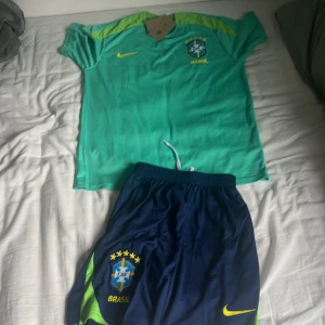 Brasilien fotbollströja och shorts från Nike - Säljer ett Brasilien fotbollskit med kortärmad grön tröja och blå shorts. Tröjan har gula detaljer och Brasilien-emblem på bröstet. Shortsen är blå med gula och gröna inslag samt Brasilien-logga och Nike-märke. Perfekt för sommarn