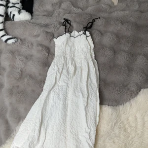 Vit långklänning med svarta knytband från H&M - Säljer en söt vit långklänning från H&M med smock upptill och svarta knytband vid axlarna. Klänningen har en lätt skrynklig struktur och svarta detaljer längs kanten. Perfekt för varma dagar!