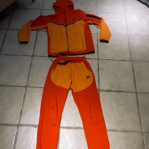 Orange träningsbyxor från Nike - Säljer ett par orange träningsbyxor från Nike med kontrasterande gula detaljer och svart Nike-logga. Byxorna har resår i midjan med snörning och är tillverkade i ett syntetmaterial.