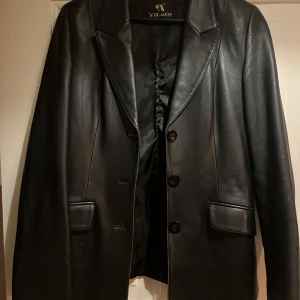 Svart skinnkavaj från YiluQi - Säljer en svart kavaj i skinnimitation från YiliQi. Kavajen har klassisk krage, tre knappar framtill och två fickor med lock. Snygg och stilren modell som passar till många olika looks.