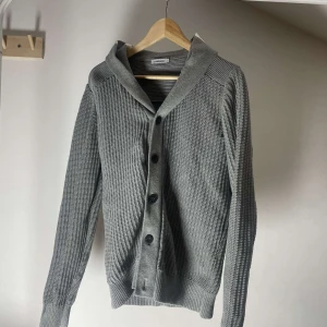 Grå stickad kofta från J.Lindeberg - Säljer en stilren grå stickad kofta från J.Lindeberg med knappar framtill och bred krage. Koftan har lång ärm och ribbade muddar. Perfekt för lager-på-lager och passar till många olika outfits.