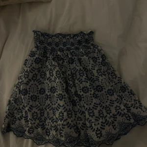 Vit kjol med blå broderade blommor - Supersöt vit kjol med blå broderade blommor och bred smock i midjan. Kjolen har vågig kant nertill och är perfekt för varma dagar. Den har en luftig och lätt känsla.