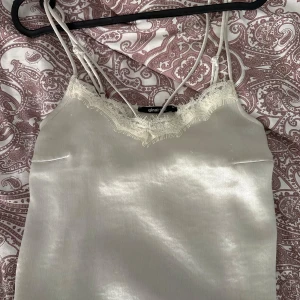 Vit satinlinne med spets Gina Tricot - Supersnyggt vitt linne från Gina Tricot i glansig satin med tunna axelband och spetsdetaljer vid v-ringningen. Perfekt att styla med jeans eller kjol för en trendig look. Sidenkänsla och romantiska vibbar .jätte liten smink fläck, vet ej om den går bort har ej provat 