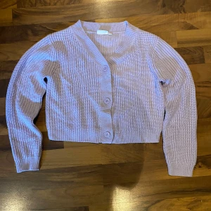 Ljuslila croppad kofta med knappar - Säljer en ljuslila stickad kofta med croppad passform och stora knappar framtill. Tröjan har långärmade ribbade muddar och bred ribbad kant vid halsen och nederdelen. Perfekt för dig som gillar mjuka och mysiga plagg med lite extra färg.