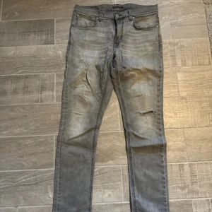 Grå jeans med slitning och rippad detalj - Säljer ett par grå jeans från Nudie Jeans med snyggt slitna detaljer och en rippad effekt på ena benet. Klassisk femficksmodell med svarta sömmar på bakfickorna. Jeansen har en rak passform och är tillverkade i bomull. I storlek 32/32