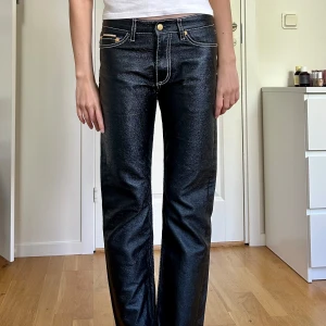 Svarta lågmidjade jeans från Eytys - Säljer ett par svarta bomullsbyxor från Eytys i glansig ”läder look” med kontrasterande vita sömmar och guldiga detaljer. Byxorna har låg midja, raka ben och klassisk femficksdesign. 