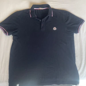 Svart Moncler pikétröja med röd/vit kant - Snygg svart pikétröja från Moncler med klassisk krage och korta ärmar. Tröjan har röd och vit kant på krage och ärmslut samt Moncler-logga på bröstet. Tillverkad i mjuk bomull som känns skön mot huden. Perfekt för dig som gillar stilrena märkesplagg.