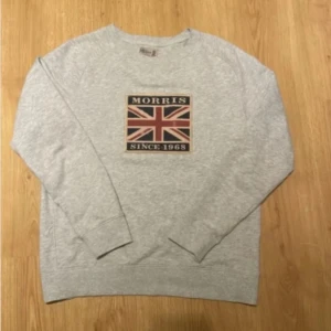 Grå Morris kofta - Snygg grå sweatshirt från Morris med Union Jack-tryck och texten 'Morris Since 1968' på bröstet. Tröjan har rund hals, långa ärmar och är gjord i mjuk bomull. Perfekt för en avslappnad och stilren look.