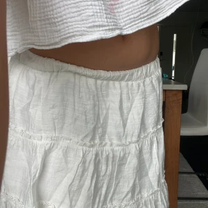 Vit volangkjol från FB Sister - Supersöt vit kortkjol från FB Sister i storlek S. Kjolen har flera volanger och ett elastiskt midjeband som gör den bekväm och lätt att bära. Perfekt för varma dagar och har en luftig känsla tack vare det tunna bomullsmaterialet.