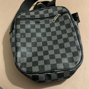 Rutig axelväska från Louis Vuitton - Säljer en snygg axelväska från Louis Vuitton med klassiskt rutigt mönster i svart och grått. Väskan har flera dragkedjefack och justerbar axelrem. Perfekt för dig som vill ha en stilren och praktisk väska med ikonisk LV-logga framtill.