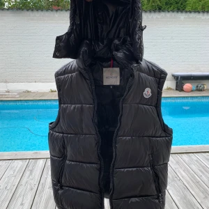 Svart dunväst från Moncler med huva (Kan diskutera pris) - Säljer en svart dunväst från Moncler med avtagbar huva och dragkedja framtill. Västen har två fickor med dragkedja och klassisk Moncler-logga på bröstet. Insidan har ett coolt serietryck och etiketter. Perfekt för lager-på-lager-stil under kyliga dagar.