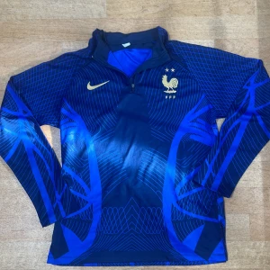 Frankrike Nike långärmad fotbollströja L - Snygg långärmad Frankrike fotbollströja från Nike i blått med guldiga detaljer och FFF-logga. Tröjan har ett unikt geometriskt mönster och är tillverkad i Dri-FIT ADV-material som andas. Perfekt för dig som vill sticka ut på planen eller på läktaren.