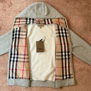 Grå Burberry hoodie med dragkedja - Säljer en grå hoodie från Burberry med klassiskt rutigt foder i beige, svart och rött. Tröjan har huva, långa ärmar och praktiska fickor framtill. Materialet är mjuk bomull och polyester, perfekt för chill dagar. Dragkedja hela vägen och snygga ribbade muddar.