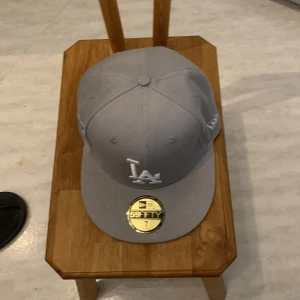 Grå LA New Era 59FIFTY keps - Snygg grå 59FIFTY keps från New Era med LA-logga framtill i vitt broderi. Klassisk flat peak och broderad MLB-logga bak. Kepsen är i storlek 7 och tillverkad i bomull. Perfekt för dig som gillar streetwear och sportig stil.