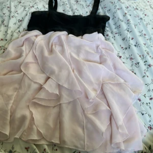 Svart och ljusrosa volangklänning H&M - Supersöt kort klänning från H&M med svart överdel och ljusrosa volangkjol. Axelbandsmodell med dragkedja i sidan för enkel påklädning. Kjolen har flera lager med mjuka volanger som ger en fluffig och drömmig look. Perfekt för dig som gillar romantisk stil!