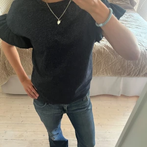 Svart glittrig topp med volangärm - Snygg svart topp från Name It med glittriga inslag och korta volangärmar. Perfekt till fest eller nyår. Passar storlek S