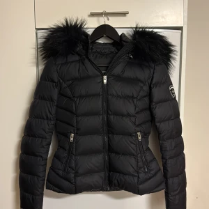 Svart dunjacka Rockandblue med fuskpäls - Snygg svart dunjacka från Rockandblue med stor fluffig fuskpäls på huvan. Jackan har quiltad design, dragkedjor i silver och Rockandblue-logga på ärmen. Figursydd modell som ger en schysst siluett och håller dig varm under kalla dagar.