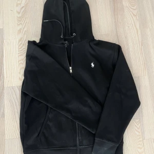 hoodie  Polo Ralph Lauren - Svart hoodie från Polo Ralph Lauren med dragkedja och huva med snörning. Klassisk logga broderad på bröstet i vitt. Tröjan har långa ärmar och ribbade muddar. Perfekt för chill dagar och enkel att matcha med jeans eller joggers. 