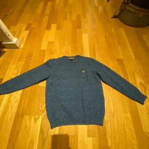 Blå stickad tröja från Lyle & Scott - Snygg blåmelerad stickad tröja från Lyle & Scott med klassisk rund hals och gul logga på bröstet. Tröjan har långärmade ribbade muddar och är perfekt för dig som gillar stilren och enkel design. Passar dig som vill ha en chill och bekväm look. Den är storlek m i barn den är som storlek 158