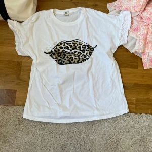 Vit t-shirt med leopardläppar River Island - Vit t-shirt från River Island med coolt tryck av leopardmönstrade läppar på bröstet. Klassisk rund hals och korta ärmar. Perfekt för dig som vill sticka ut med en trendig och lekfull detalj. Materialet känns mjukt och skönt mot huden.