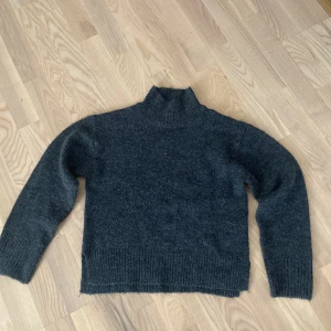 Mörkgrå stickad polotröja - Mysig mörkgrå stickad polotröja med hög krage och långa ärmar. Tröjan har en enkel, klassisk design med ribbade muddar vid ärmslut och nederkant. Perfekt för kyliga dagar när du vill vara både bekväm och snygg. Bra för hösten och vintern 