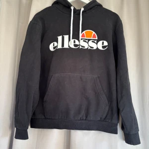 Svart Ellesse hoodie med logga - Svart hoodie från Ellesse i storlek EU 38. Tröjan har vit Ellesse-logga med orange detalj på bröstet, vit snörning i huvan och en stor magficka. Mjuk insida och ribbade muddar vid ärmslut och nederkant. 