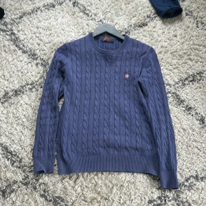 Blå kabelstickad tröja från Morris - Säljer en blå kabelstickad tröja från Morris i 100% pima bomull. Tröjan har rund halsringning, ribbade muddar och en liten broderad logga på bröstet. Perfekt för dig som gillar klassisk och stilren look.