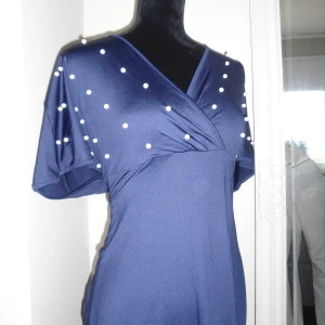 Marinblå omlottklänning med pärlor - Snygg marinblå omlottklänning med korta ärmar och v-ringad hals. Överdelen är dekorerad med vita pärlor som ger en cool och unik look. Klänningen har en mjuk, följsam passform.