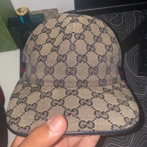 Gucci Navy keps med GG-mönster - Säljer en beige keps från Gucci med ikoniskt GG-mönster i mörkblått. Kepsen har justerbar rem bak i skinn och snygga detaljer med blå och röd rand på sidorna. Materialet är bomull och skinn, perfekt för dig som vill ha en lyxig och trendig look.