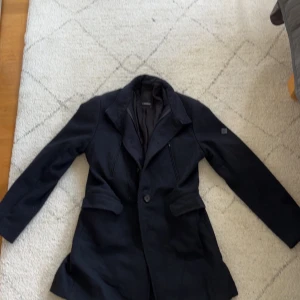 Svart rock från J.Lindeberg, stl 48 - Stilren svart rock från J.Lindeberg med klassisk krage, knäppning framtill och snygga fickdetaljer. Jackan har ett diskret logomärke på ärmen och bälte med knappar bak. Perfekt för dig som vill ha en clean och tidlös look. Den är även lite nopprig skulle nog behöva en tvätt eller trimmas ( jag kan trimma om du inte har en egen trimmer). Syns inte så bra bilderna men jackan har en dragkedja under knapparna. Kom med frågor och prisförslag.