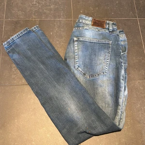 Blå slitna jeans med färgstänk - Säljer ett par blå jeans med slitningar och målarfärg för en cool, avslappnad look. Jeansen har klassisk femficksdesign, normal passform och är tillverkade i jeansmaterial. Perfekta för dig som gillar en streetig stil.