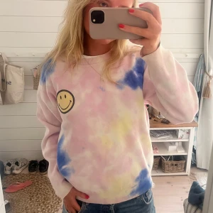 Sandro tröja - Superfin pastellfärgad tie-dye tröja med smiley-märke på bröstet. Mjuka färger i rosa, blått och gult. Rund halsringning och långa ärmar. Perfekt för dig som gillar färg och vill sticka ut lite extra.