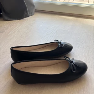 Svarta ballerinaskor med rosett - Klassiska svarta ballerinaskor med rund tå och söt liten rosett framtill. Skorna har en platt sula och är fodrade med beige tyg invändigt. Perfekta för en stilren och enkel look.