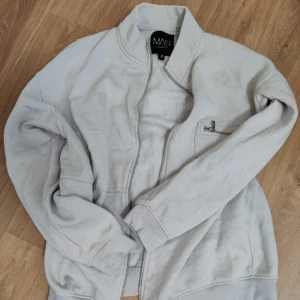 Ljusgrå ziptröja från MAN - Säljer en ljusgrå ziptröja från MAN med ståkrage och hel dragkedja framtill. Tröjan har en ficka med dragkedja på ena ärmen och ribbade muddar vid ärmslut och nederkant. Perfekt för chill dagar och enkel att matcha med jeans eller joggers.