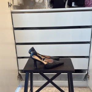 Svarta pumps med spetsig tå i skinn 37 - Snygga svarta pumps i äkta skinn med spetsig tå och smal klack. Skorna har en elegant design med diskreta sömdetaljer och öppen häl med rem och spänne. Perfekta för dig som vill ha en klassisk och stilren look.