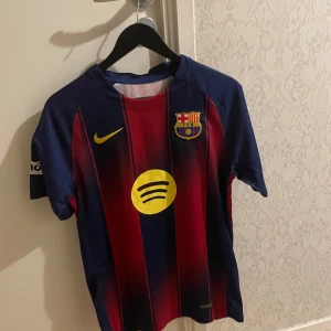FC Barcelona Lamine Yamal #10 matchtröja - Säljer en officiell FC Barcelona matchtröja från Nike med Lamine Yamal och nummer 10 på ryggen. Tröjan har klassiska blå och röda vertikala ränder, gula detaljer och klubbmärke på bröstet. Materialet är lätt och andas, perfekt för fotboll eller häng med kompisar.