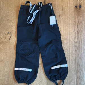 Nya Svarta täckbyxor Polarn O. Pyret 122 - Svarta skalbyxor från Polarn O. Pyret med justerbara (och avtagbara) hängslen. Vind och vattentäta Resår i midjan och benslut. Storlek 122, helt nya.