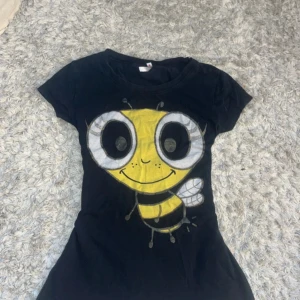 Svart t-shirt med bi-tryck - Svart t-shirt med stort, färgglatt tryck av en tecknad bi på framsidan. T-shirten har rund hals och korta ärmar. Materialet känns mjukt och lätt, perfekt för dig som gillar roliga motiv och vill sticka ut lite extra.