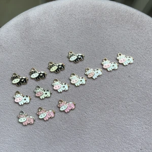Söta berlocker med ko-motiv - Samling med gulliga berlocker i form av små kor i olika färger: svart, vit och rosa. Perfekta att använda till halsband, armband eller örhängen. Berlockerna har detaljer i guld och emalj, och några har en 3D-form med orange mule. Skapa egna smycken med dessa unika och charmiga motiv!