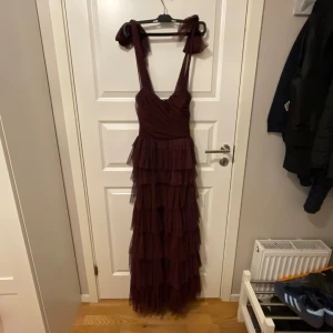 Vinröd långklänning med volanger - Säljer en vinröd långklänning med flera lager av volanger i tyll. Klänningen har breda axelband med knyt och en omlottliknande överdel. Perfekt för dig som vill sticka ut med en romantisk och elegant look.