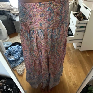 Färgglad långkjol med paisleymönster - Säljer en bohemisk långkjol med paisleyinspirerat mönster i rosa, blått och gult. Kjolen har smock i midjan och är tillverkad i ett lätt, flowy material som passar perfekt för varma dagar. Volangdetaljer längst ner ger extra rörelse och stil.